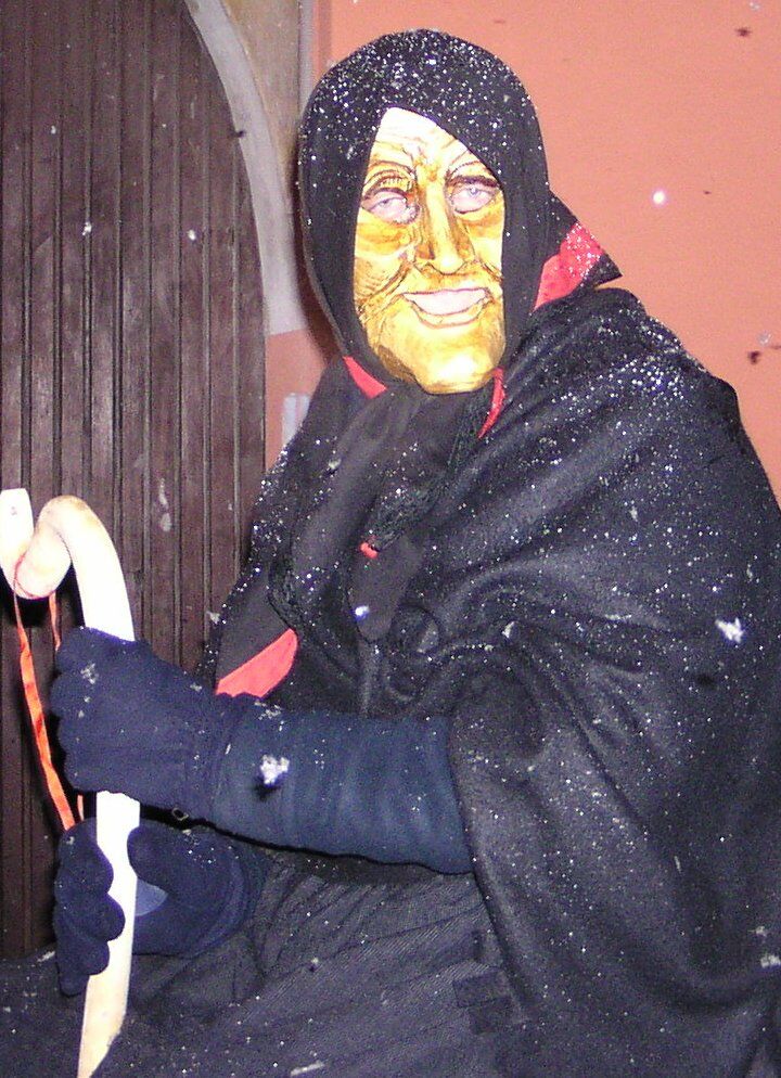 Figura mascherata da vecchia con scialle nero e volto giallo