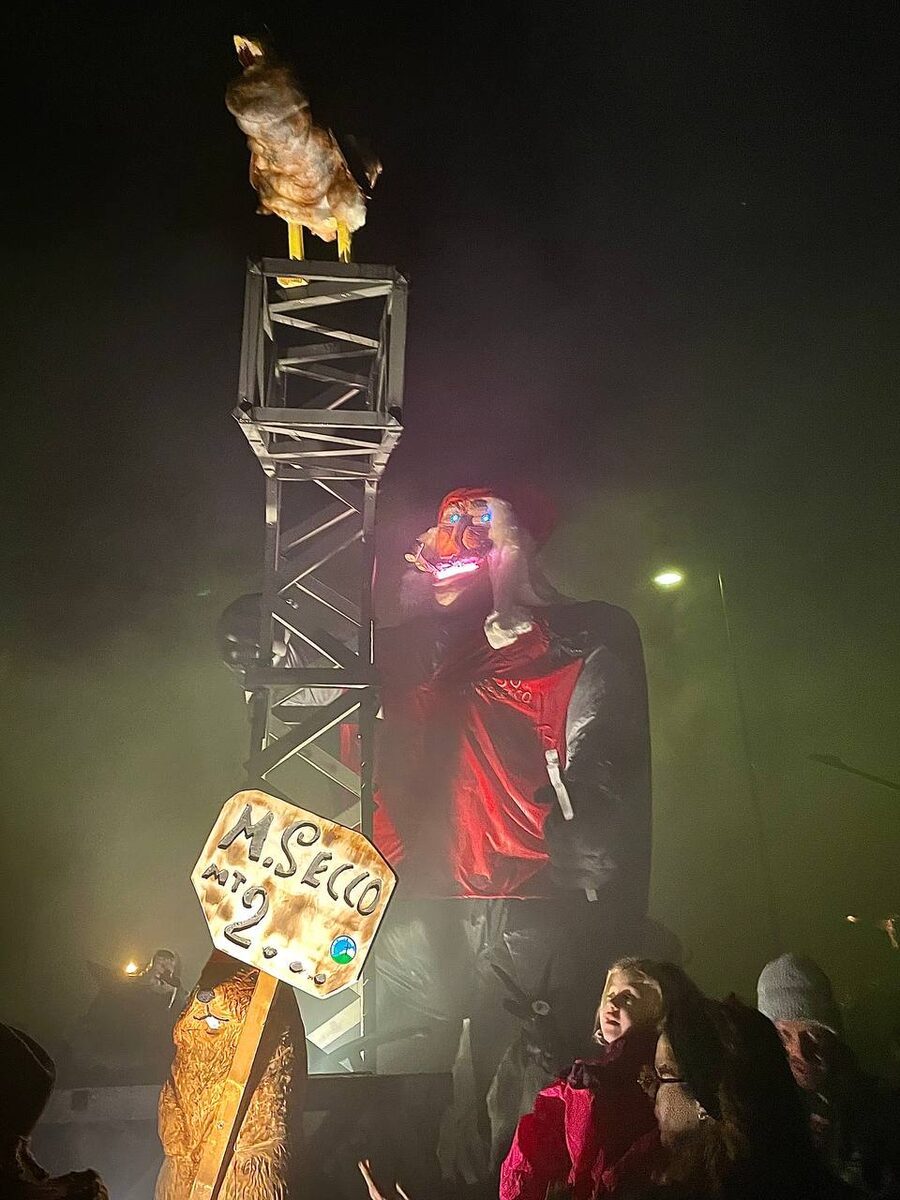 Il fantoccio del Zenerù illuminato di notte, con giacca rossa, accanto a un traliccio