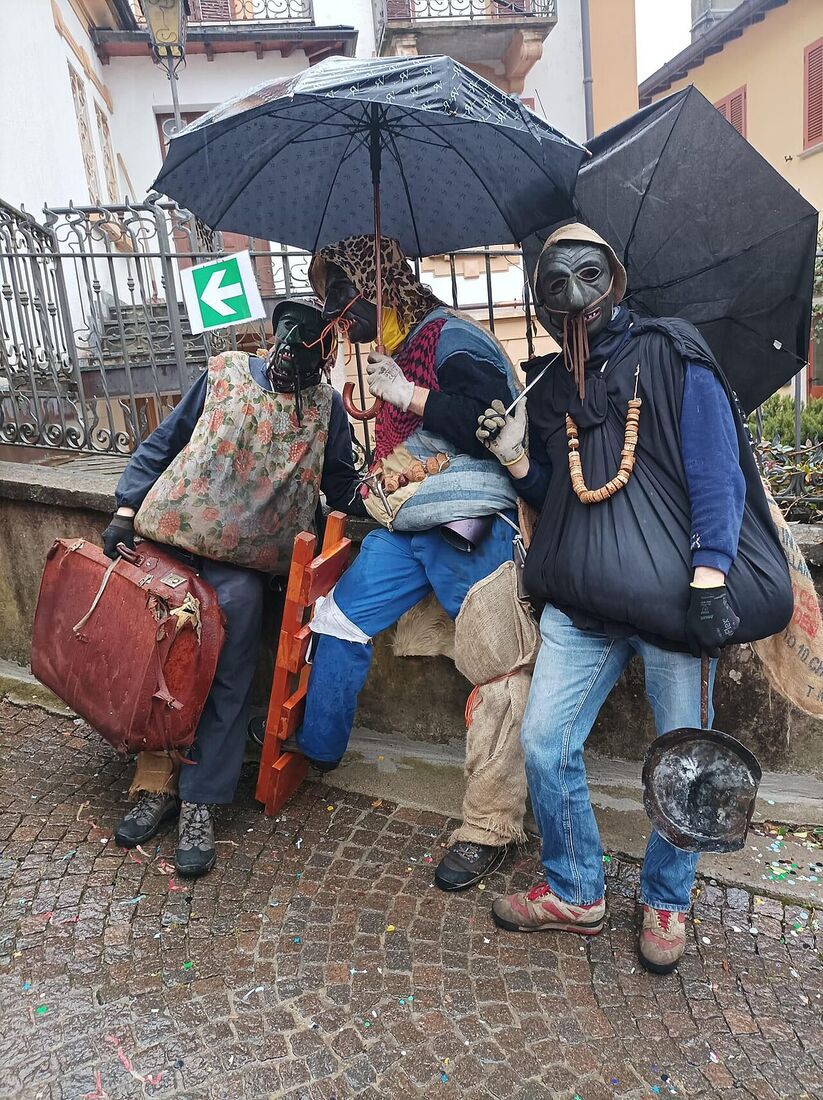 Maschere di Brutti del Carnevale di Schignano sedute con valigie e ombrelli